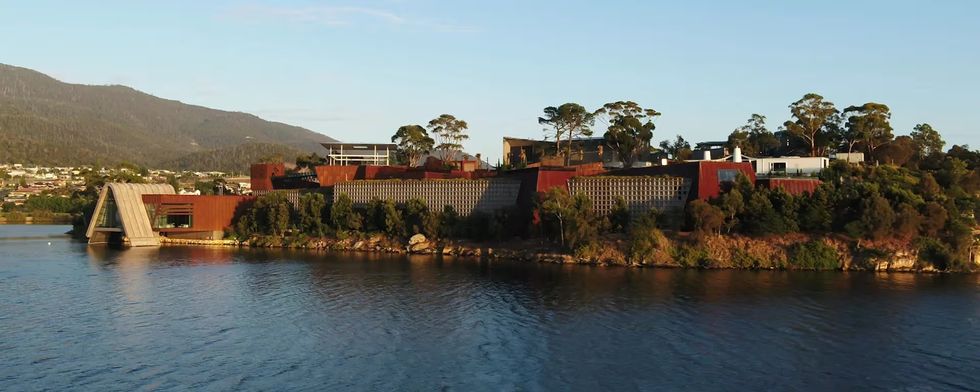 Modern building complex called Mona by a riverside, surrounded by hills and trees.