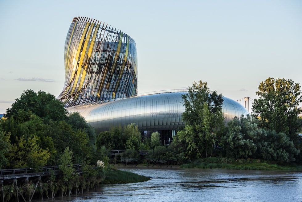 Modern curved building, Cit\u00e9 du Vin, by a river surrounded by green trees under a clear sky.