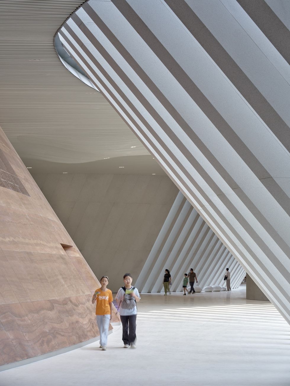 Modern interior of \u200bShenzhen Natural History Museum with slanted beams; two people walk, sunlit striped shadows on the floor.