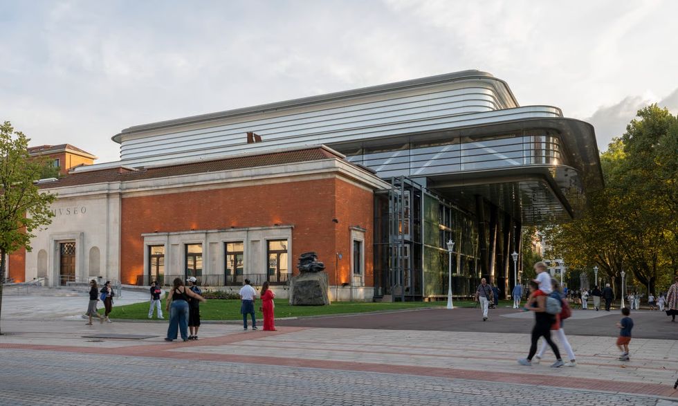 Modern museum exterior with brick facade and large glass extension, people walking nearby.