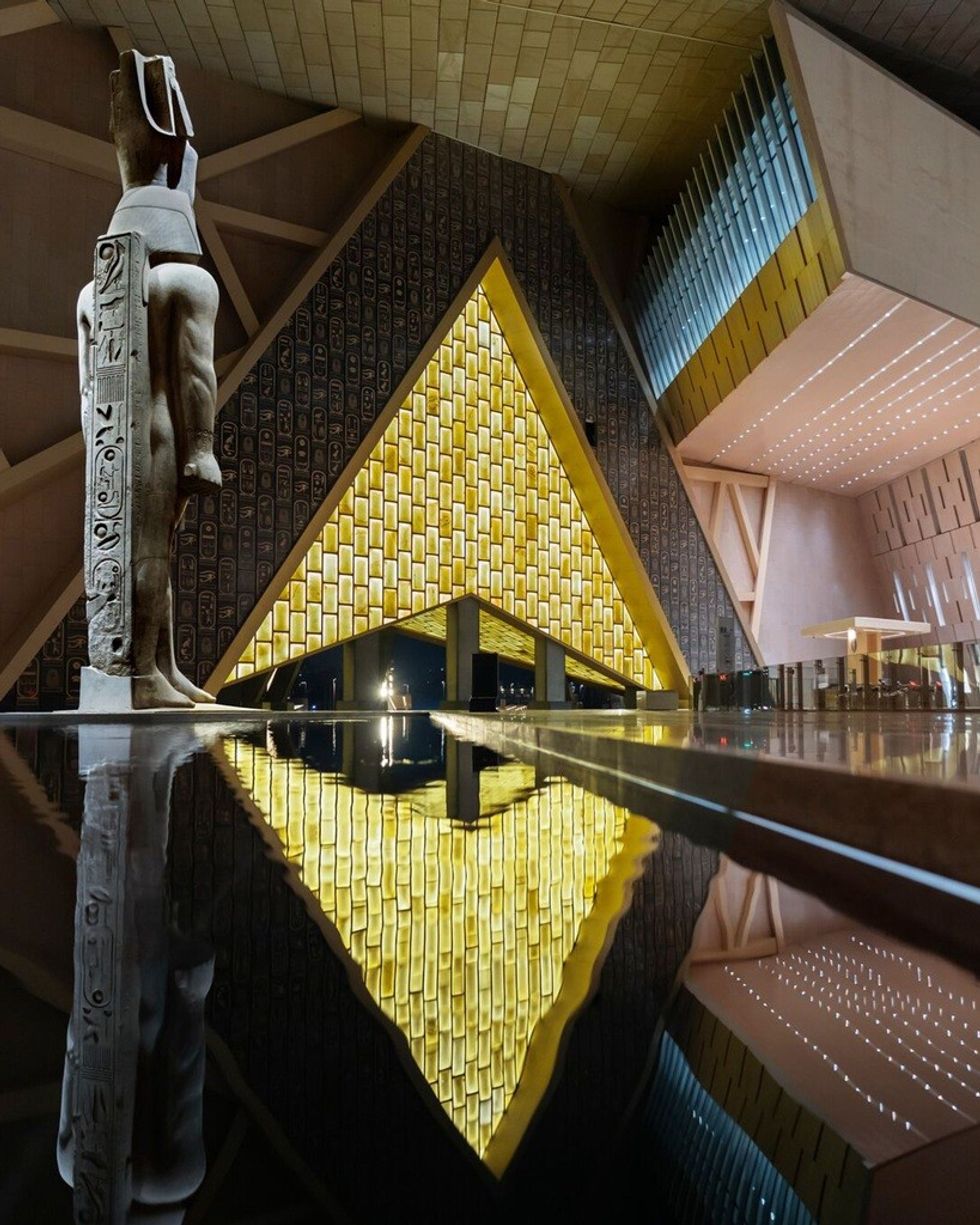 Modern museum interior with geometric architecture, statue, and water reflection.