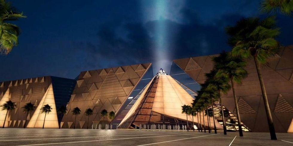Modern pyramid structure at the Grand Egyptian Museum in Giza, illuminated at night with a light beam and palm trees surrounding it.
