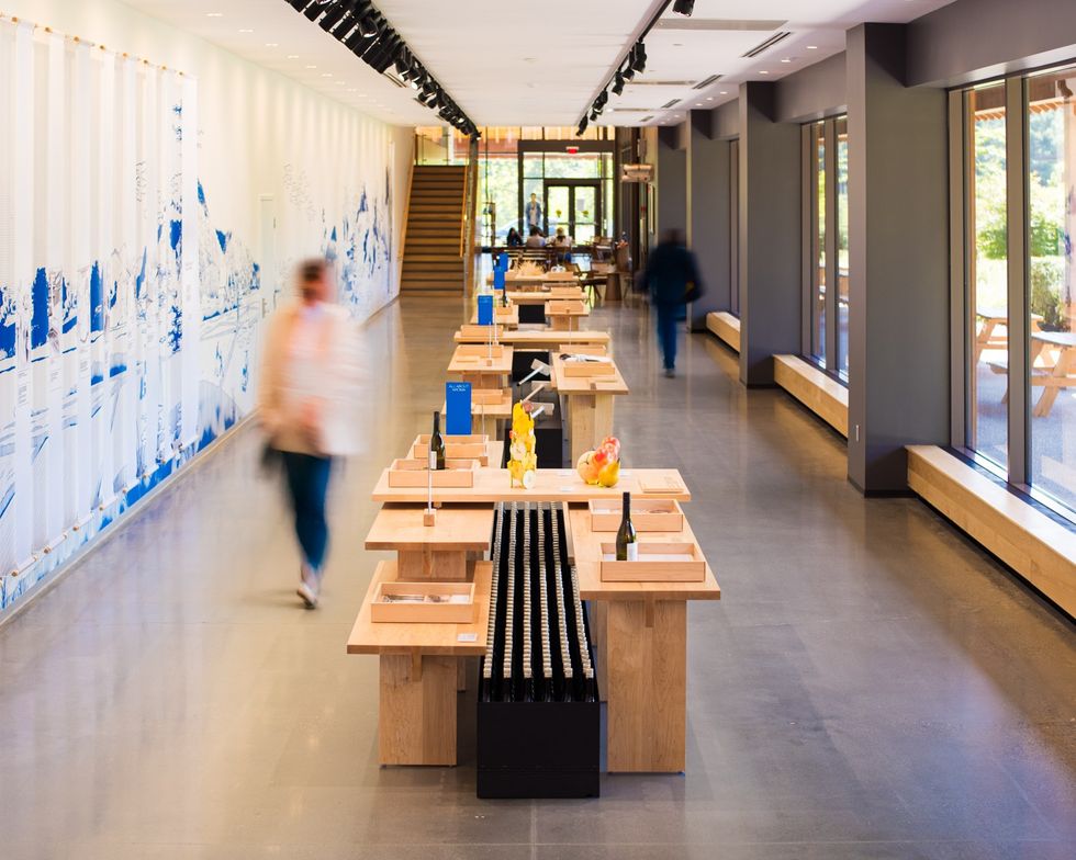 Modern tasting room with wooden tables and blurred visitors, large windows on the right.