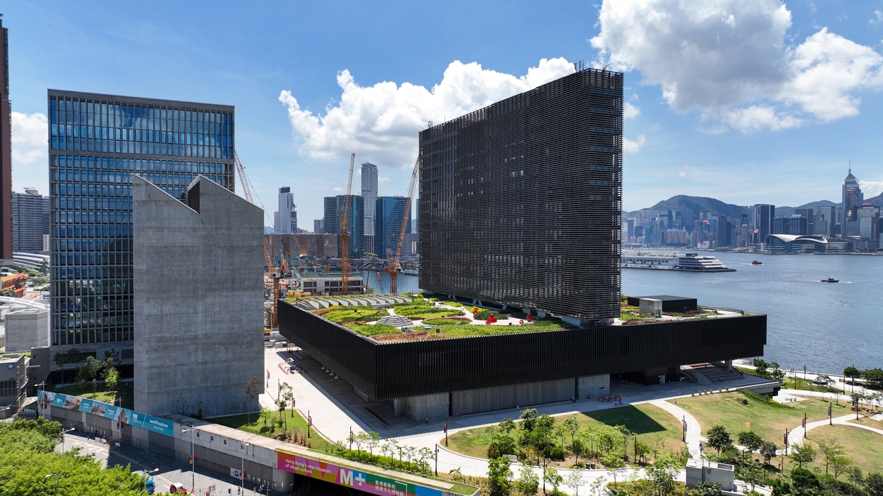 Modern waterfront M+ museum with a rooftop garden, harbor, and city skyline beneath a bright, cloudy sky.