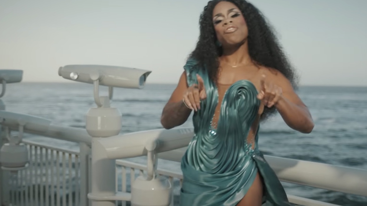Monét X Change in a flowing teal dress posing near oceanfront railings.