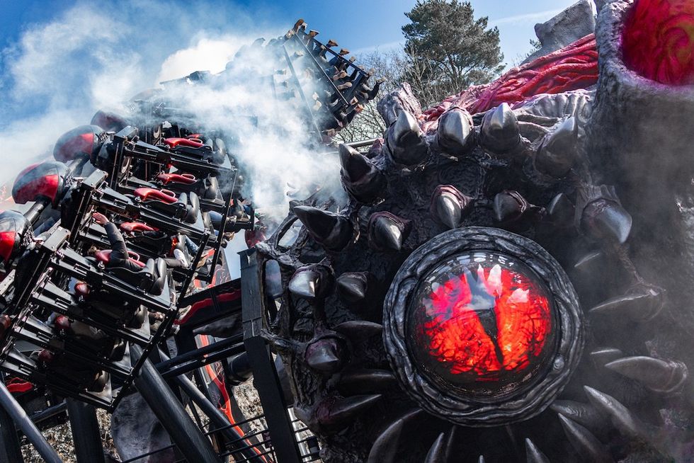 Nemesis Reborn roller coaster with smoke and a giant, red, eye-like structure with spikes, against a blue sky.