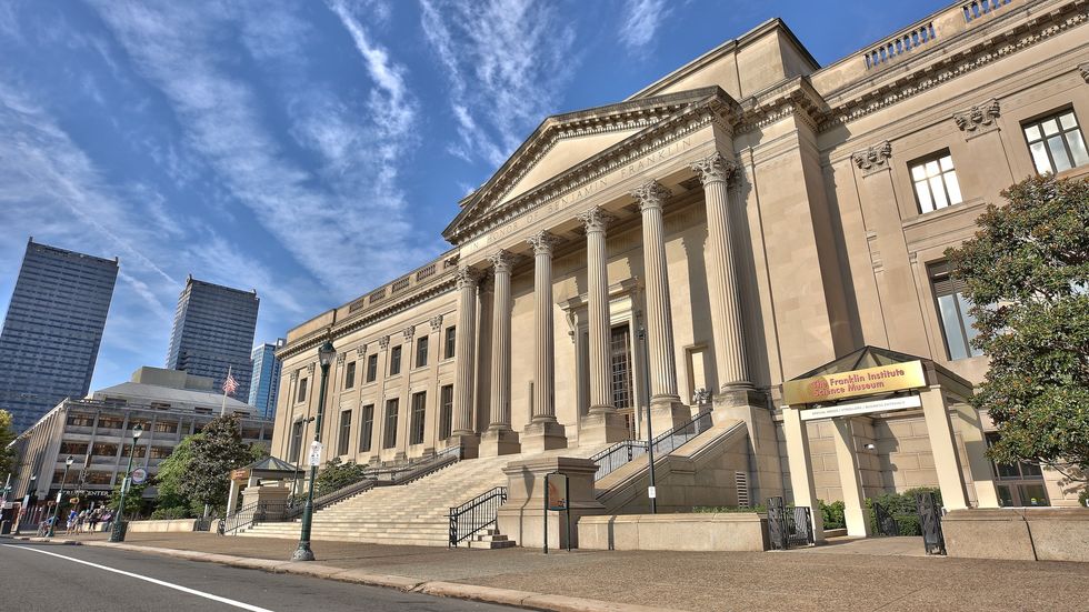 Neoclassical building with columns; Franklin Institute entrance; modern skyscrapers in background.