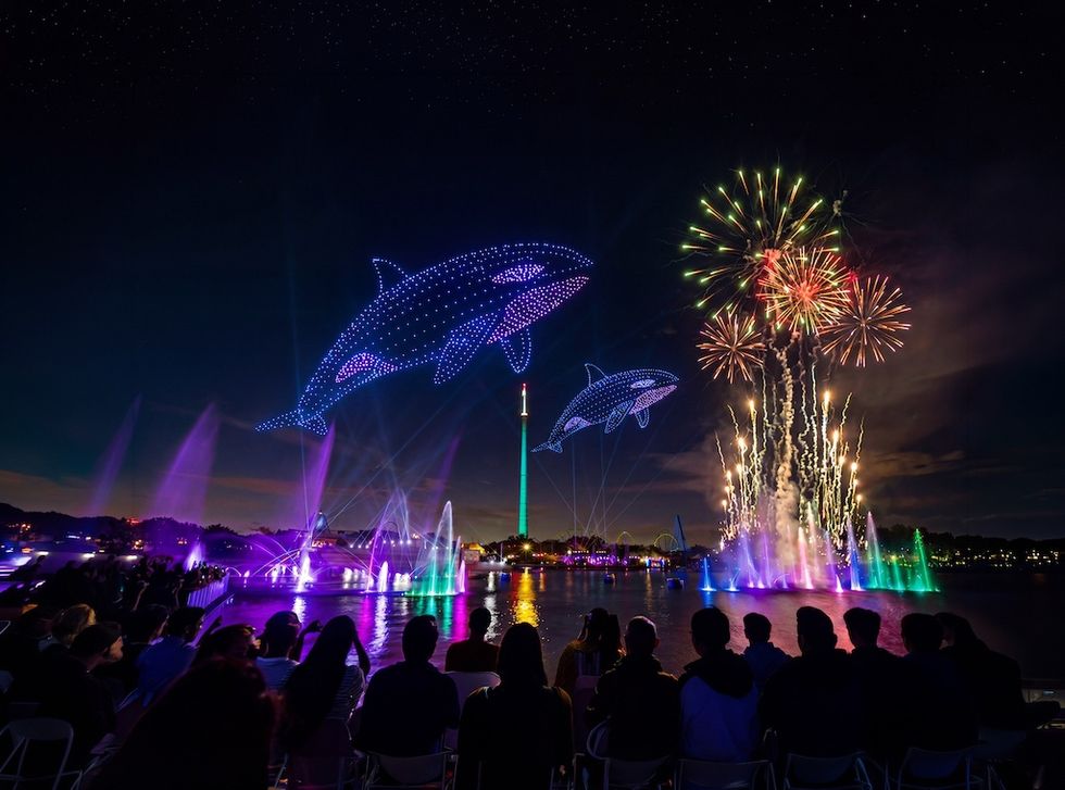 Night sky lit by fireworks, drone whales, colorful fountains, and silhouetted crowd near water.