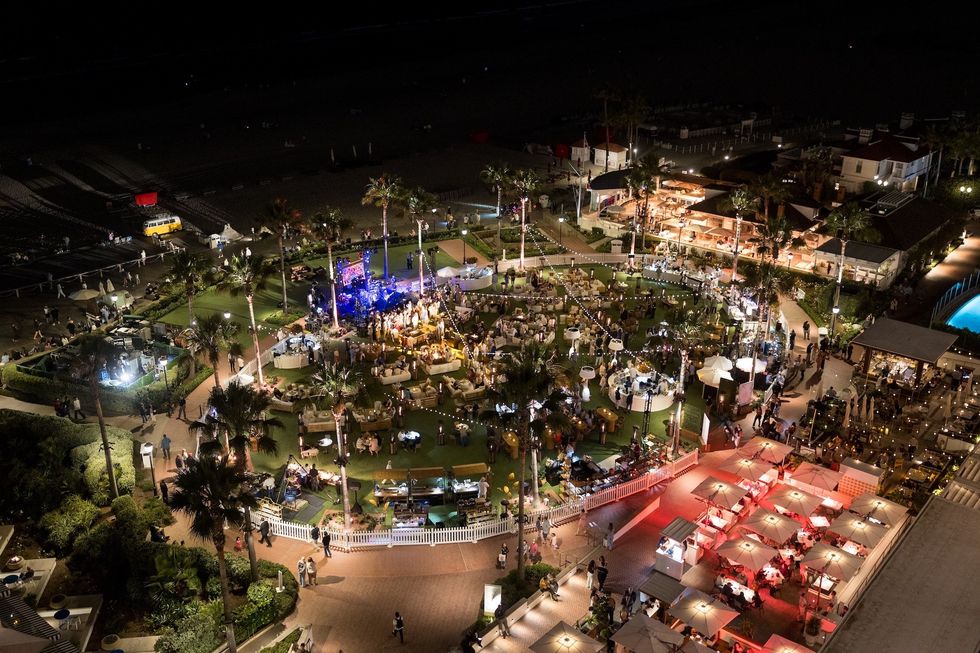 Nighttime outdoor event with lights, tables, and crowds on a well-lit lawn near the coast.