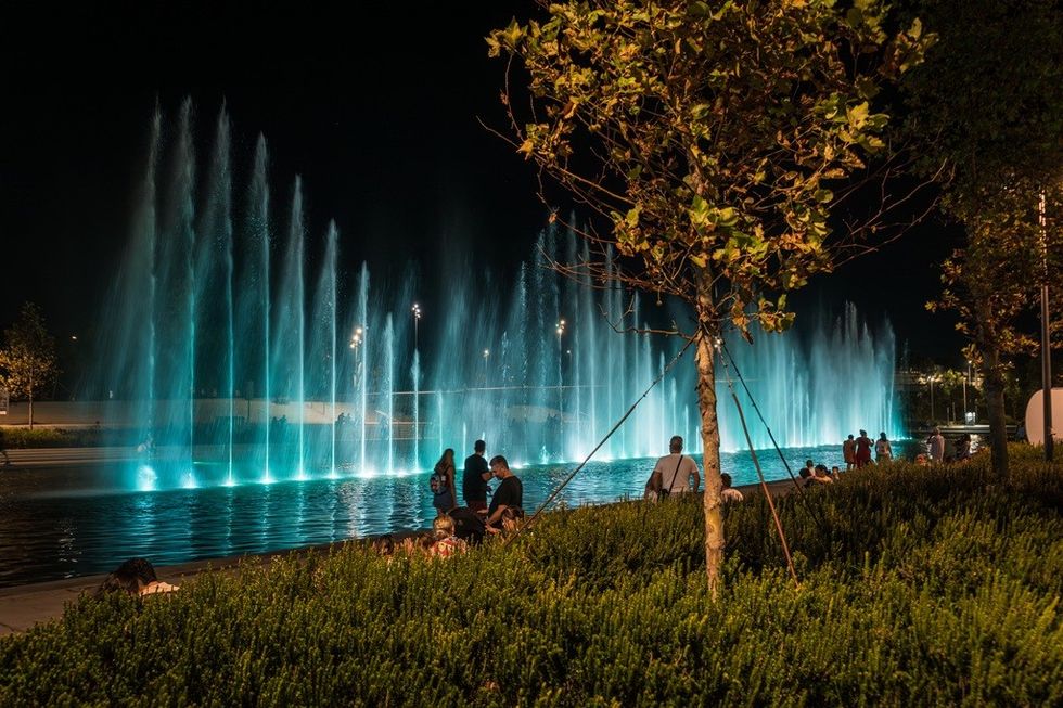 Nighttime park scene with illuminated fountains and people gathered around.