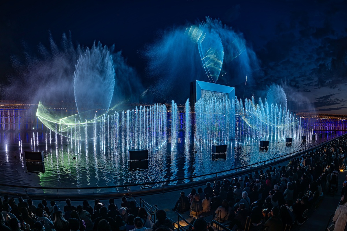 Nighttime water fountain show with blue lights and holographic projections, crowd watching.