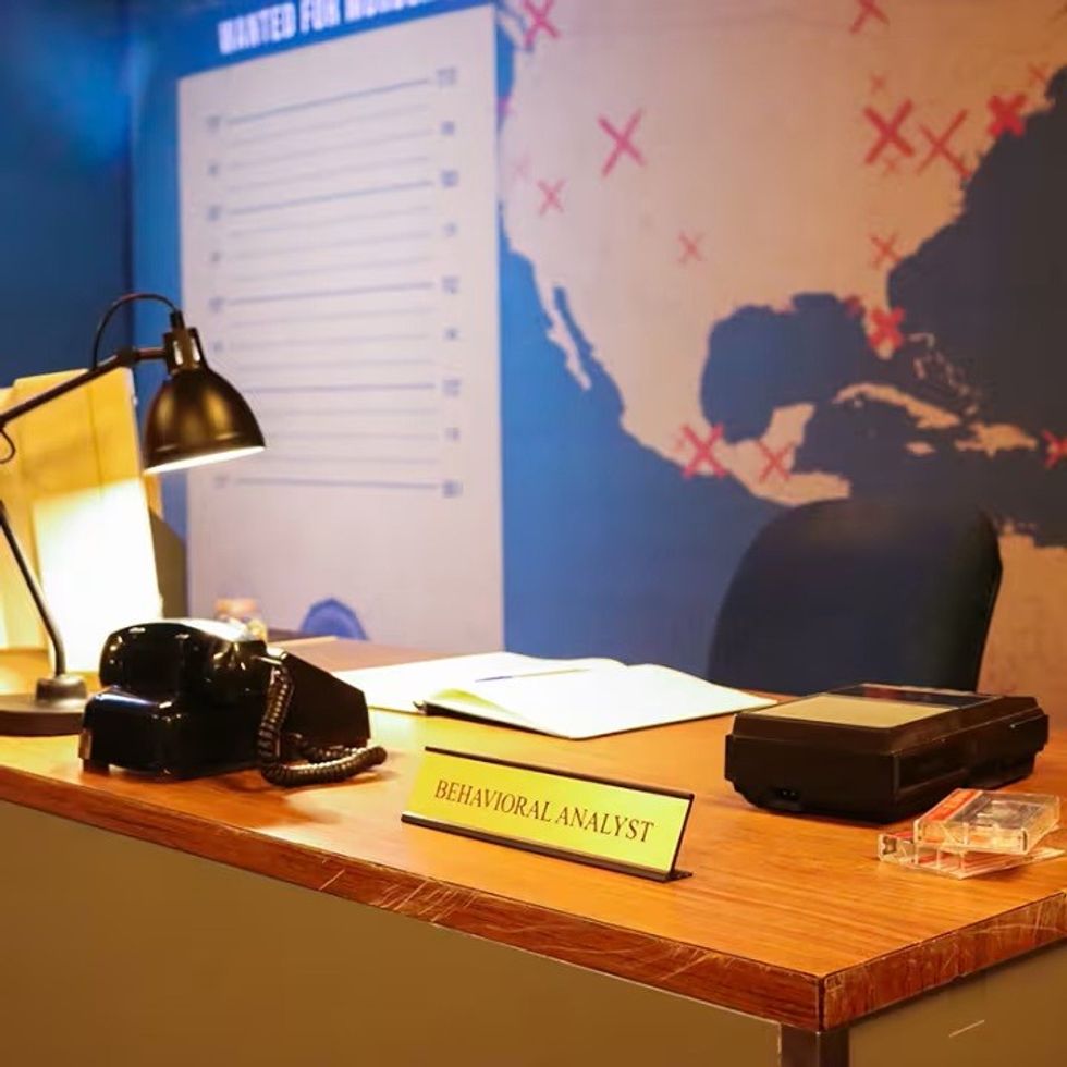 Office desk with "Behavioral Analyst" sign, lamp, phone, and map with marked locations.