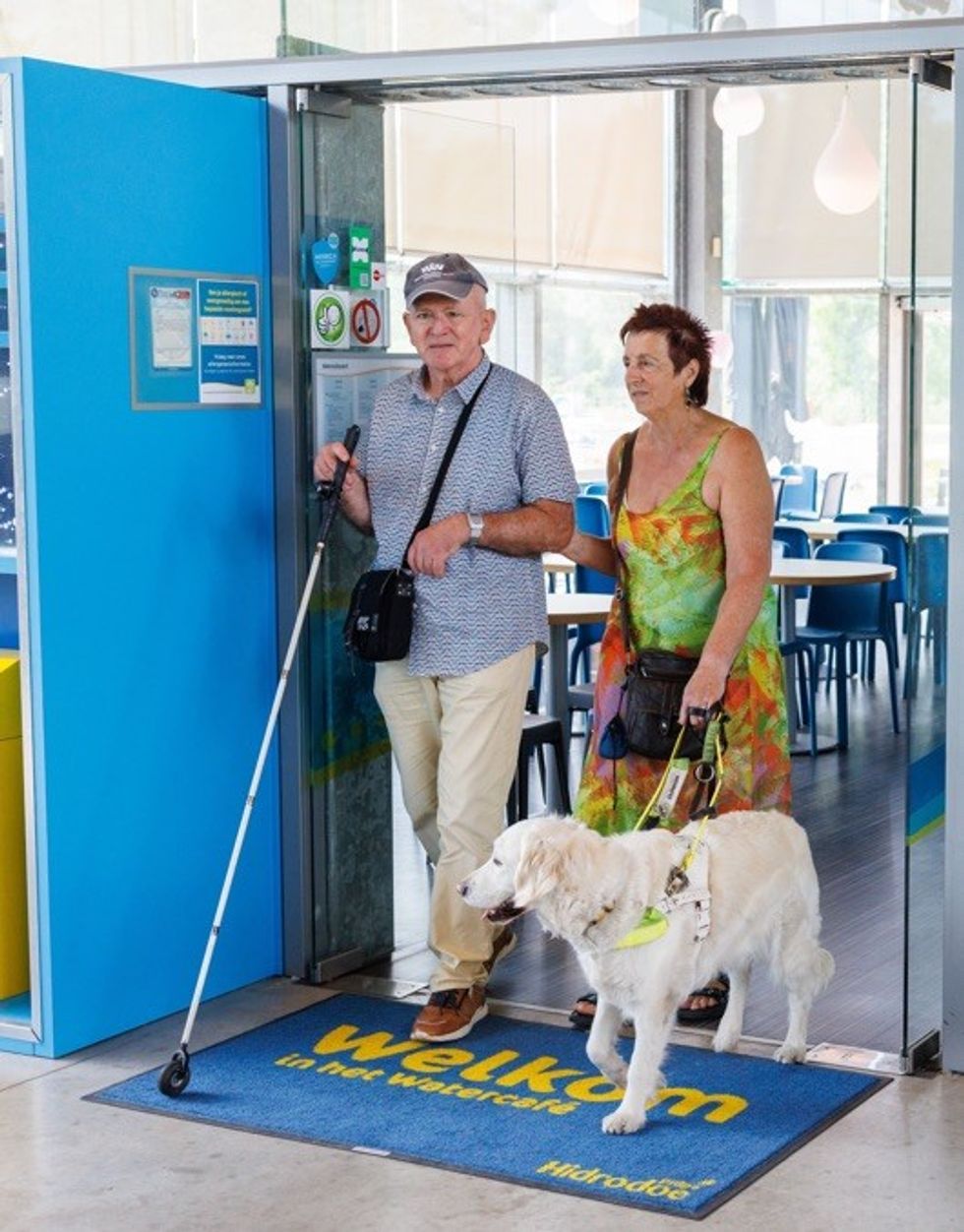Older couple with a guide dog entering a cafe, woman supporting the man with a white cane.