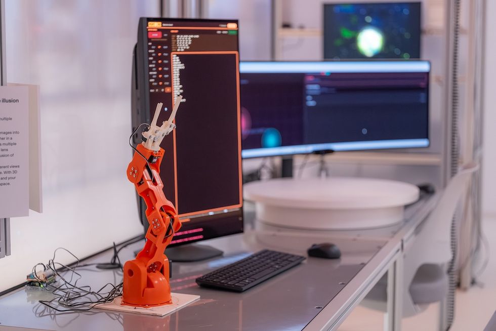 Orange robotic arm beside computer monitors displaying code in a tech lab.