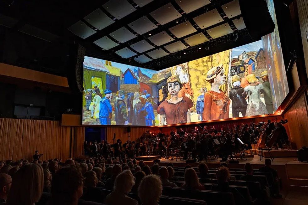 Orchestra playing beneath a large, vibrant painting projected in a concert hall.