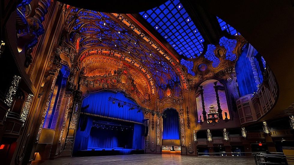 Ornate theater stage with blue curtains, intricate ceiling, warm lighting, and detailed decor.