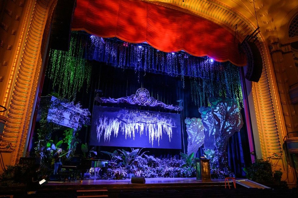 Ornate theater stage with lush foliage, red curtains, and colorful lighting.