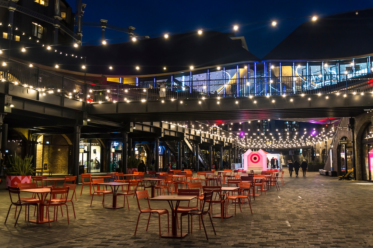 Outdoor dining courtyard at night, with empty tables, string lights, and a modern glass-fronted building.