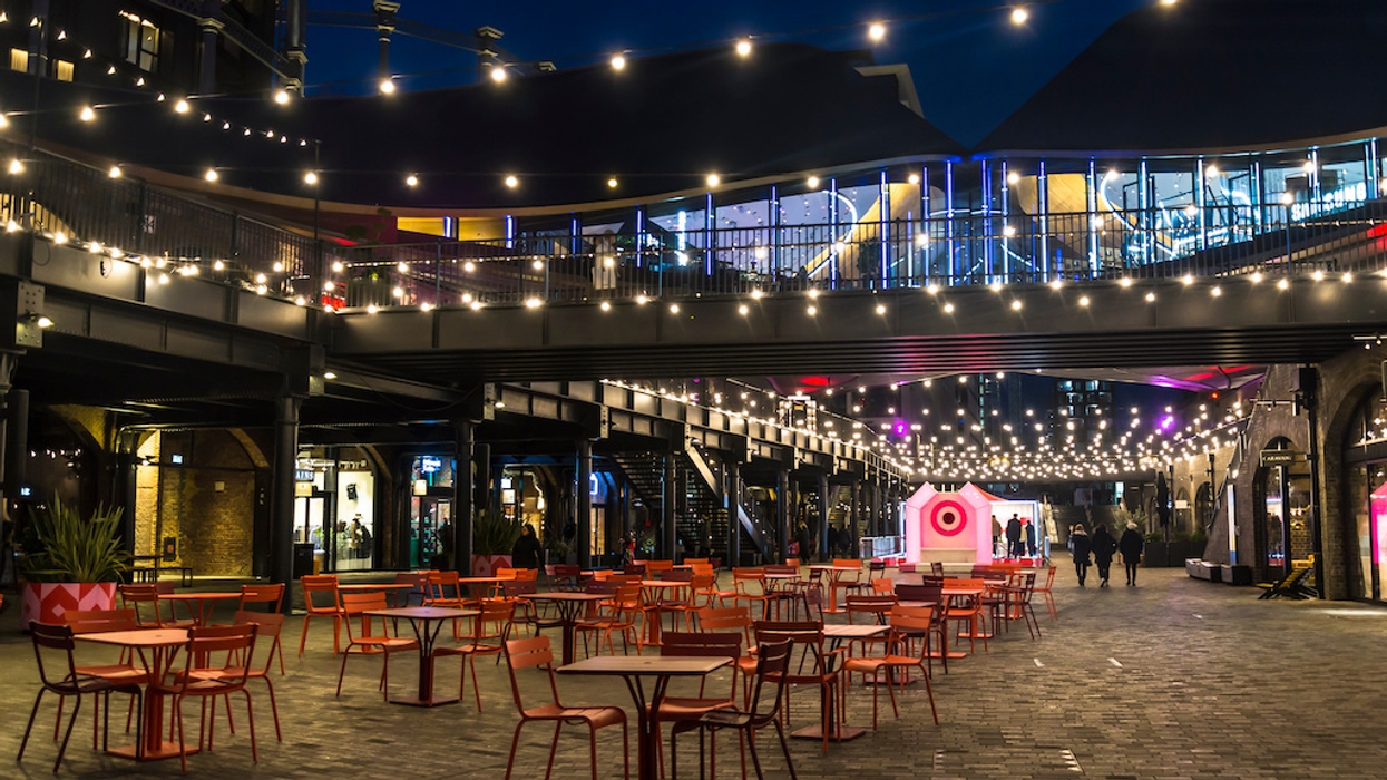 Outdoor dining courtyard at night, with empty tables, string lights, and a modern glass-fronted building.