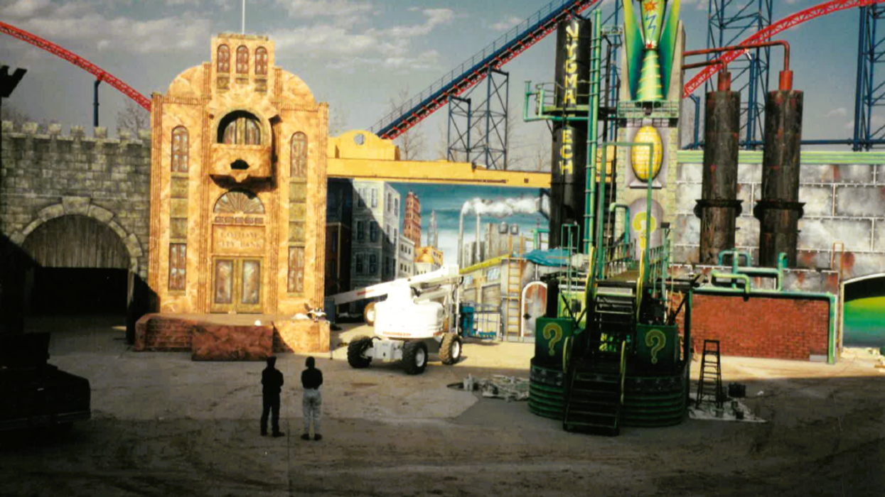 Outdoor movie set with colorful props and industrial structures; roller coasters in background.