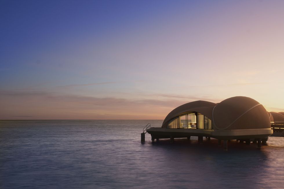Overwater villa at sunset with a calm ocean backdrop and a clear sky.