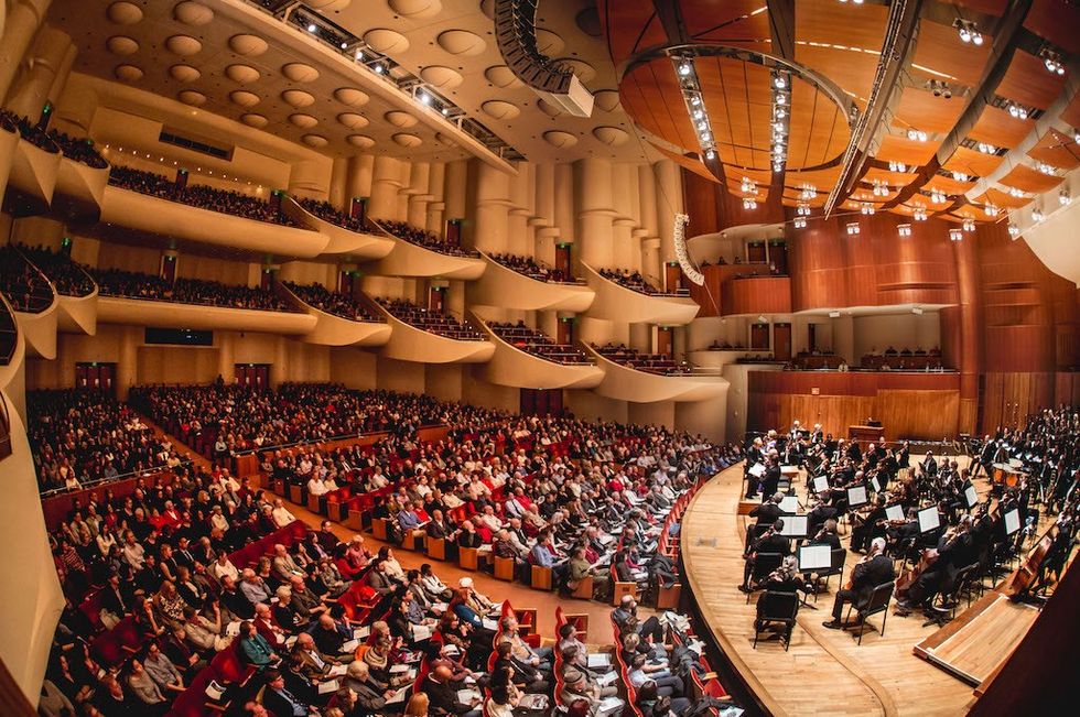 Packed concert hall with orchestra performing, audience seated in modern, multi-tiered venue.