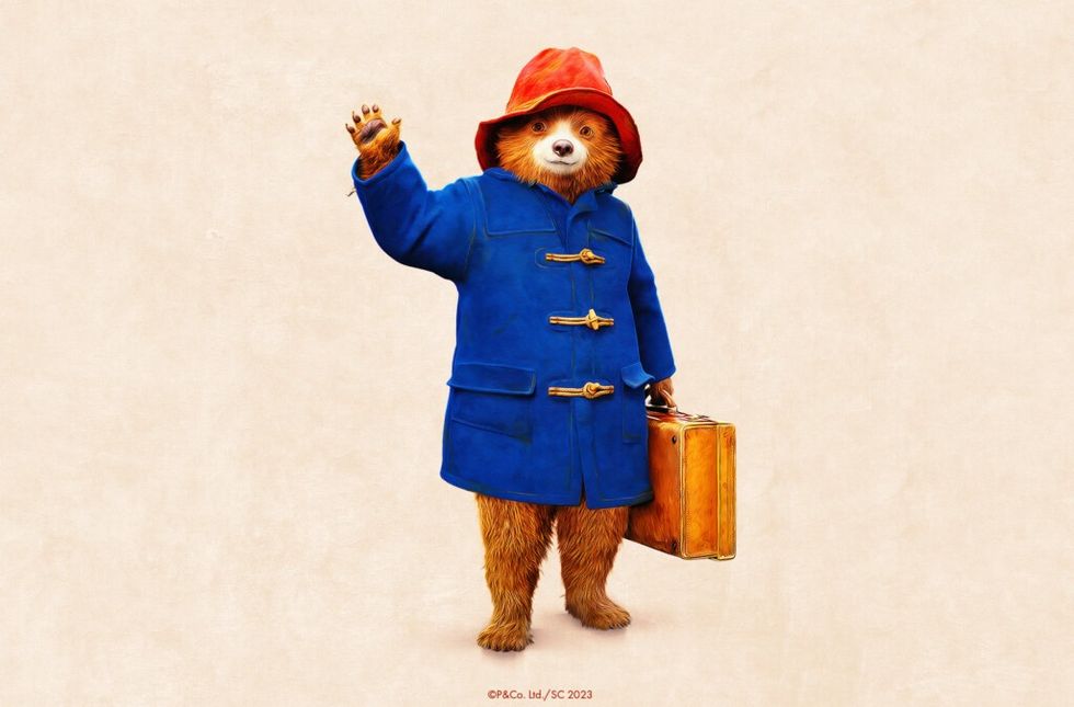 Paddington Bear in a blue coat and red hat waving and holding a suitcase.