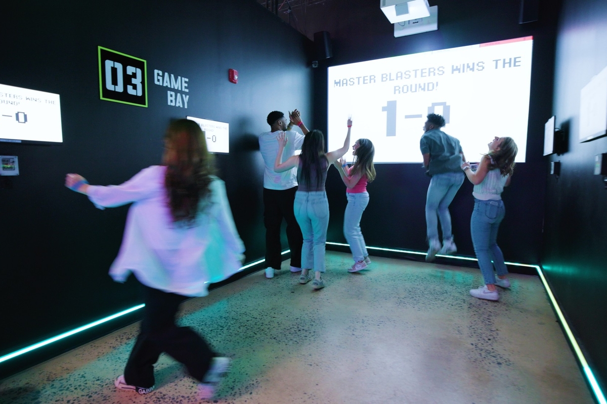 People celebrating in a gaming room with a scoreboard reading "Master Blasters Wins 1-0".