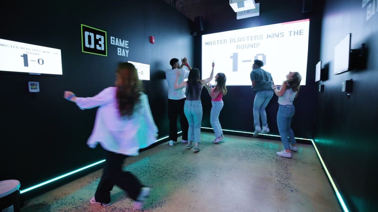 People celebrating in a gaming room with a scoreboard reading "Master Blasters Wins 1-0".