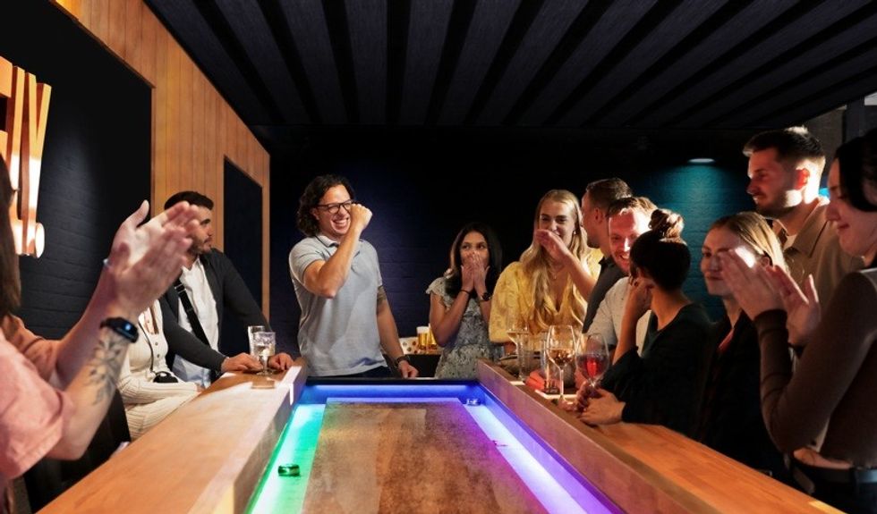 People cheer around a bar with a lighted shuffleboard game.