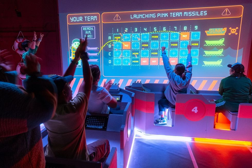 People cheering while playing an interactive missile-themed game with a large screen display, Squid Game Experience