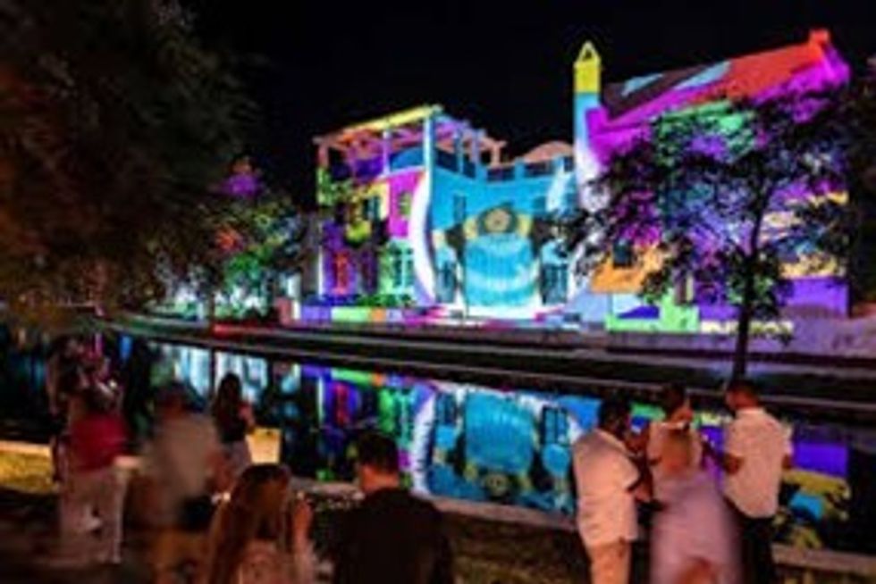 People enjoy vibrant light projections on a building by a waterway at night.