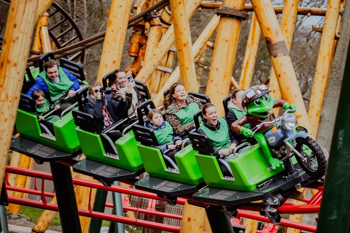People enjoying a ride on a green roller coaster with a frog mascot in front.