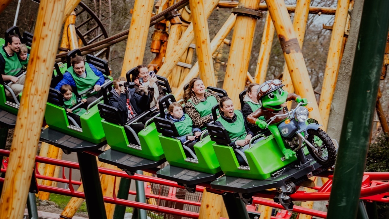 People enjoying a ride on a green roller coaster with a frog mascot in front.