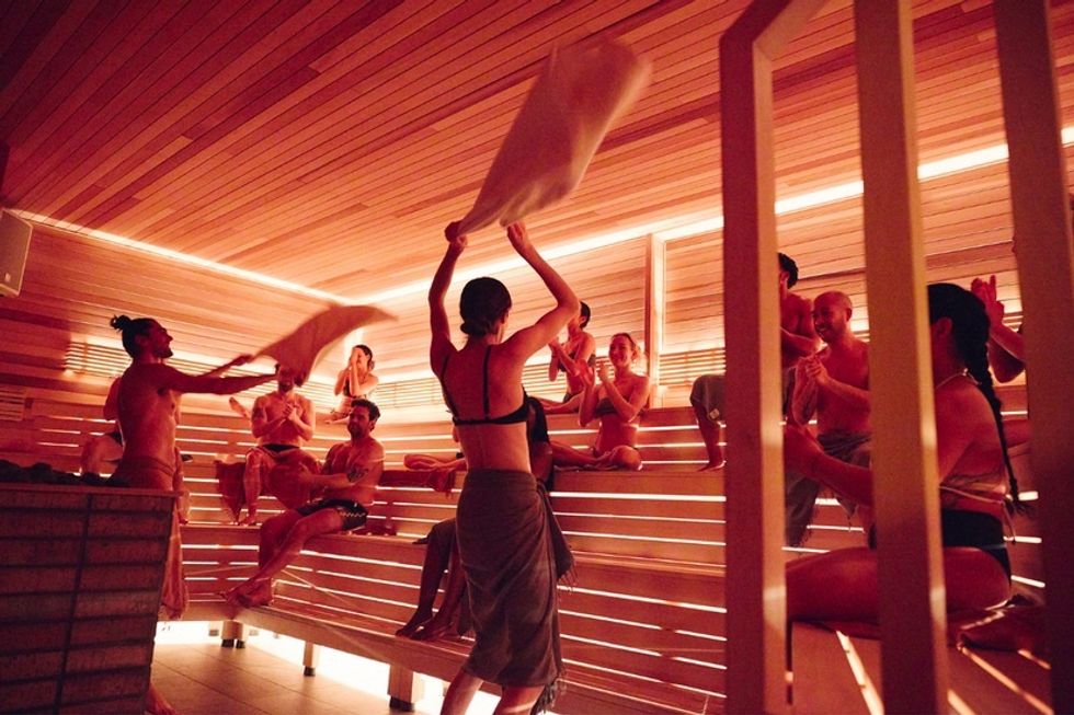 People enjoying sauna, waving towels, and sitting on wooden benches in a warm, ambient lit room.