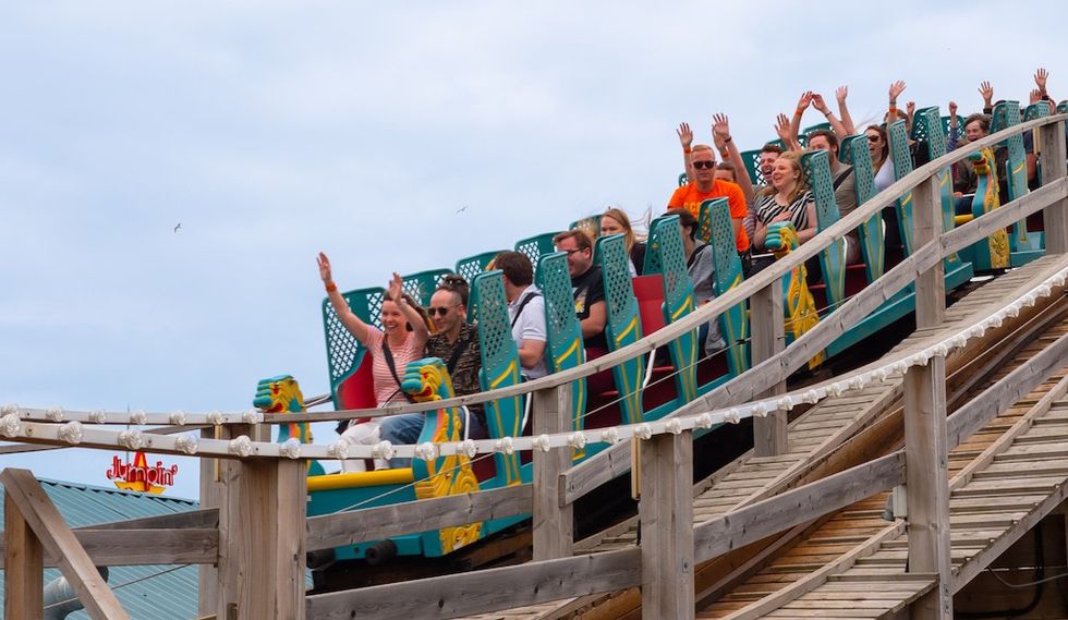 People excitedly riding a wooden roller coaster with hands in the air under a cloudy sky.