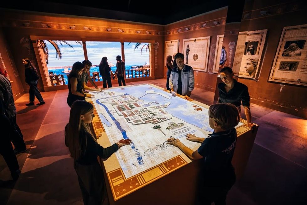 People gather around an interactive table in a historical exhibit room with large windows.