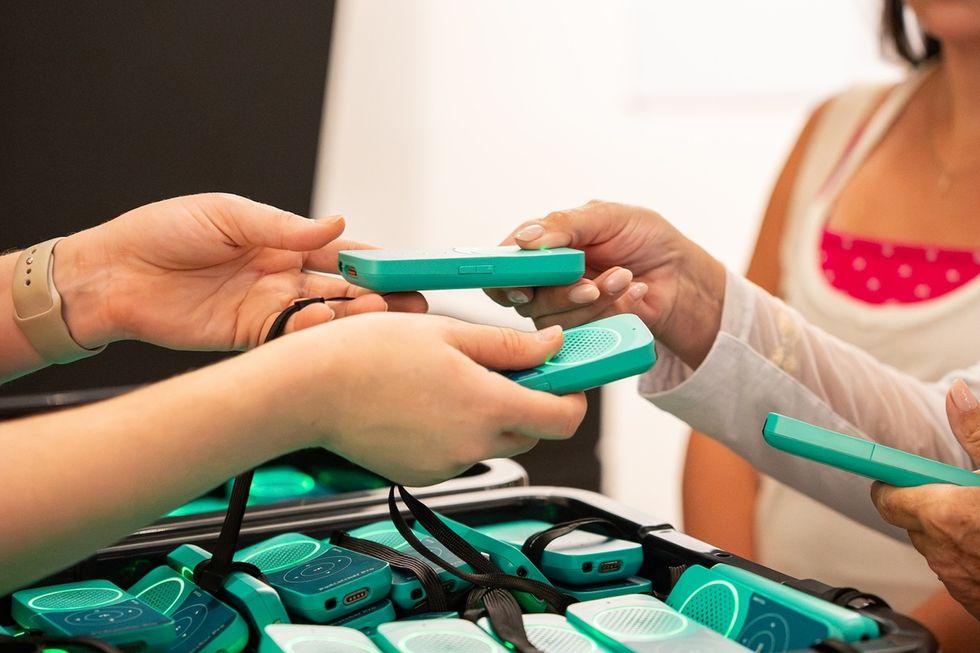 People handle teal Guide-ID audio tour devices above a case filled with matching units.
