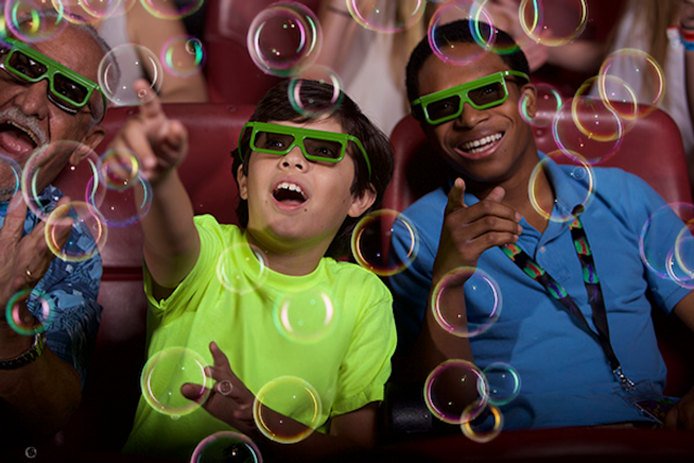 People in 3D glasses enjoy bubbles in a theater, reaching and smiling.