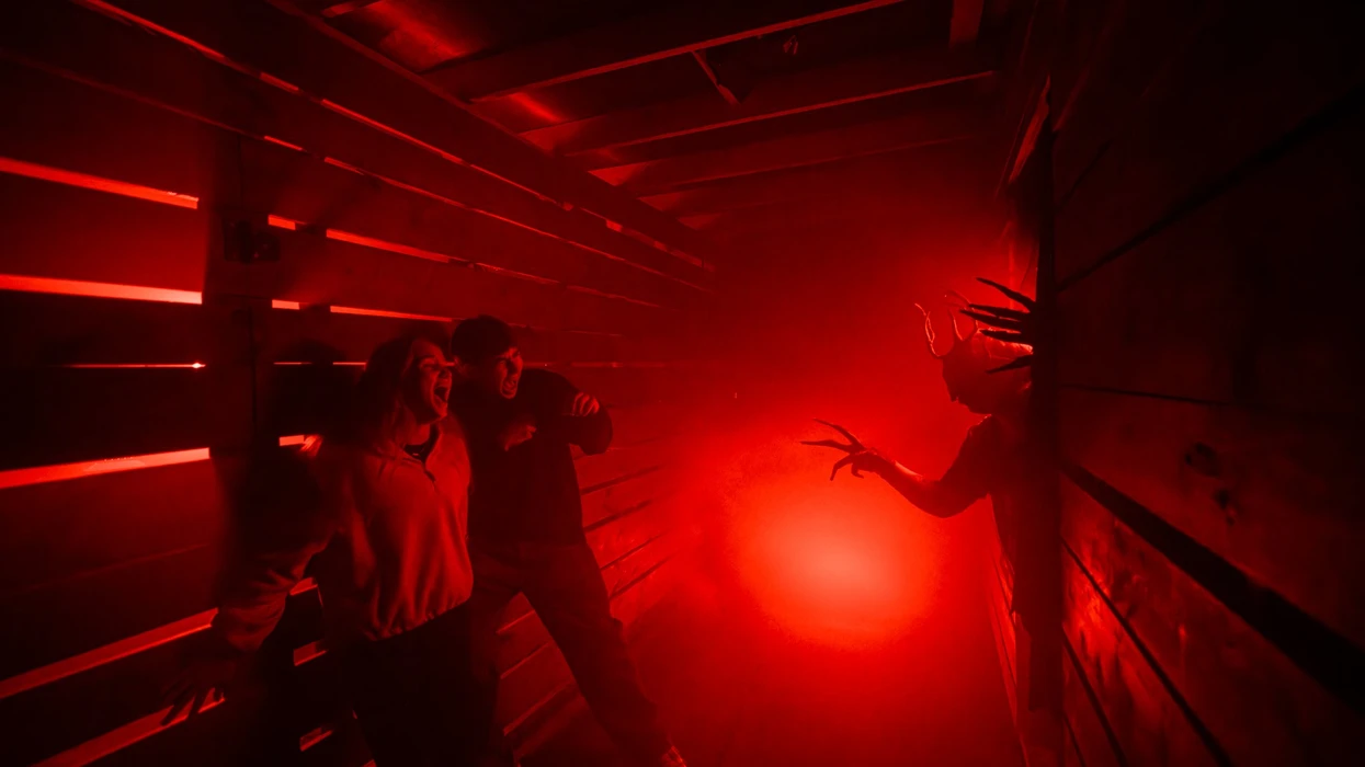 People in a haunted house corridor scream at a creature with long claws in red lighting.