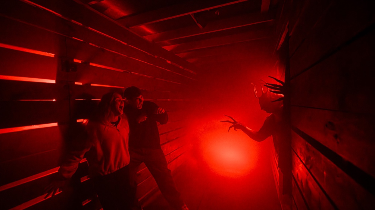 People in a haunted house corridor scream at a creature with long claws in red lighting.
