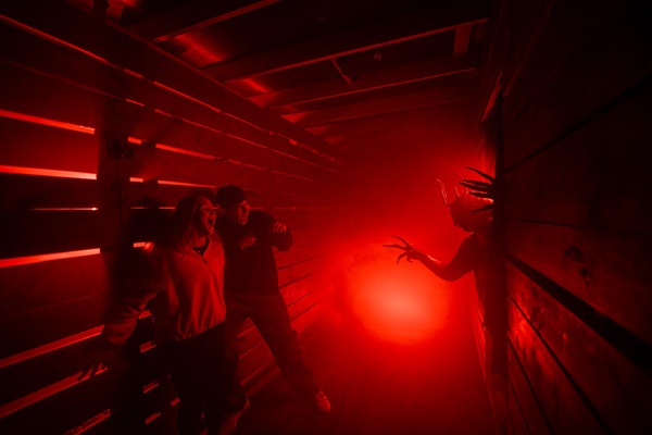 People in a haunted house corridor scream at a creature with long claws in red lighting.