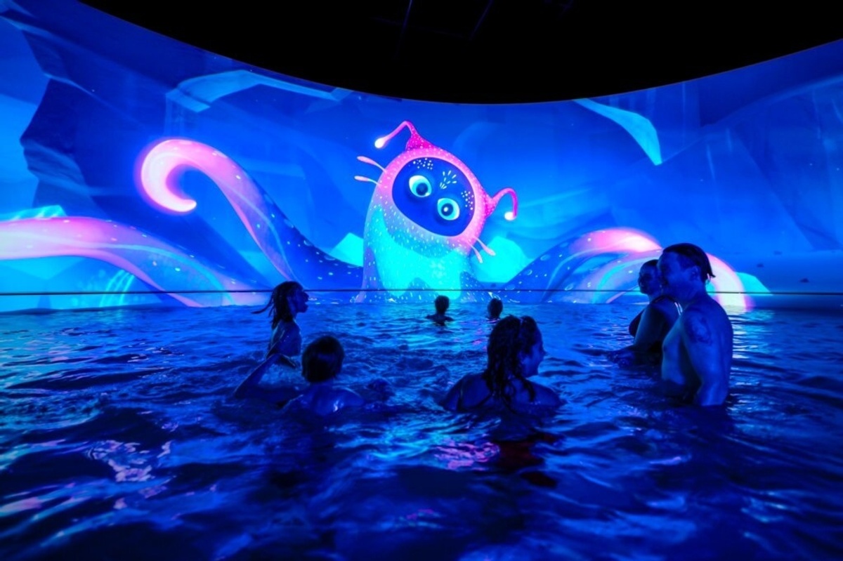 People in a pool at Aquascope in France, watching a bright, animated creature on a large screen.