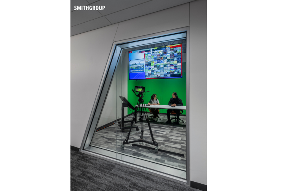 People in a studio with cameras and a wall of screens visible through a glass window.