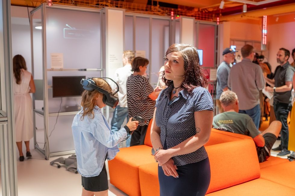 People in a tech exhibit; child wearing VR headset, woman in foreground, vibrant setting.