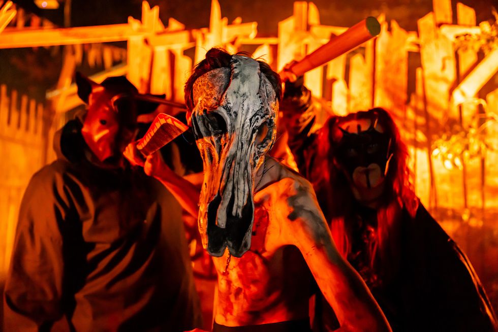 People in eerie masks, holding props, in a dimly lit, spooky setting.