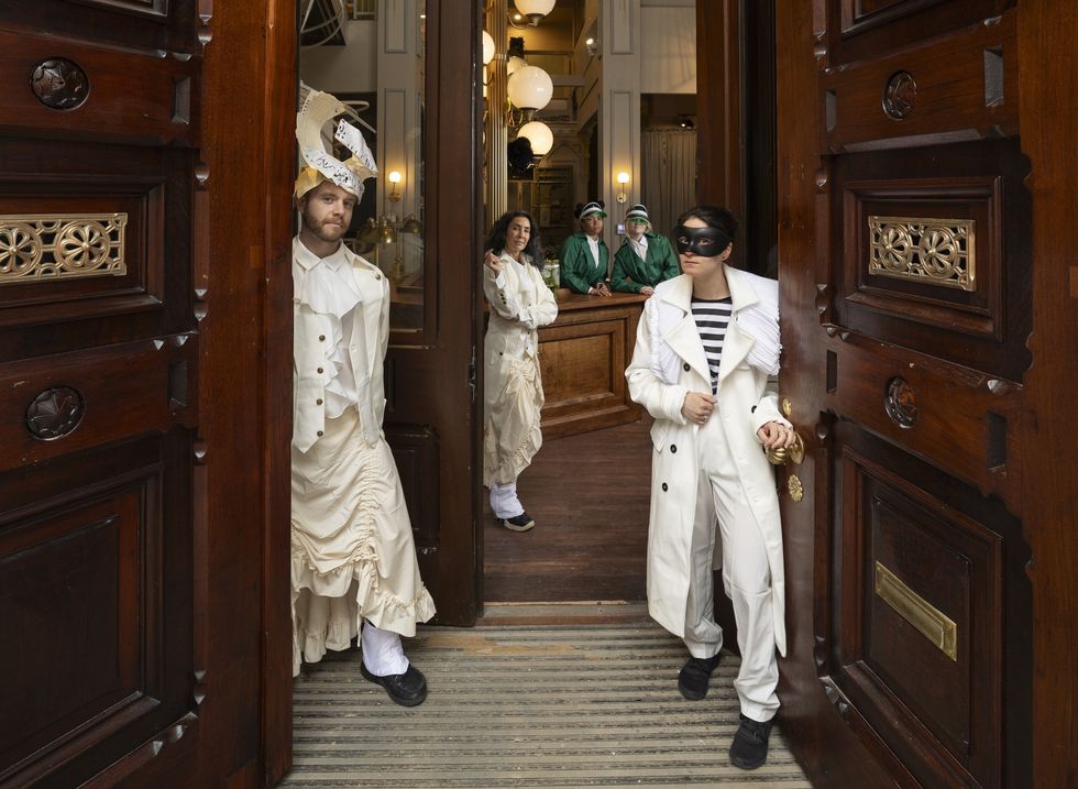 People in elaborate costumes and a mask stand by open ornate wooden doors at the Ministry of Awe.