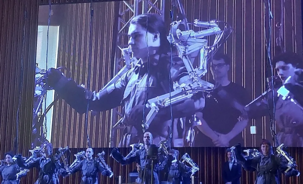 People in exoskeleton suits perform on stage with a large screen backdrop at Ecsite 2024