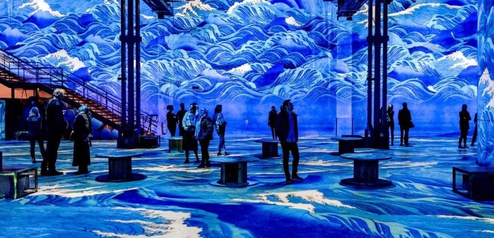 People in immersive art exhibit with swirling blue wave projections on walls and floor.