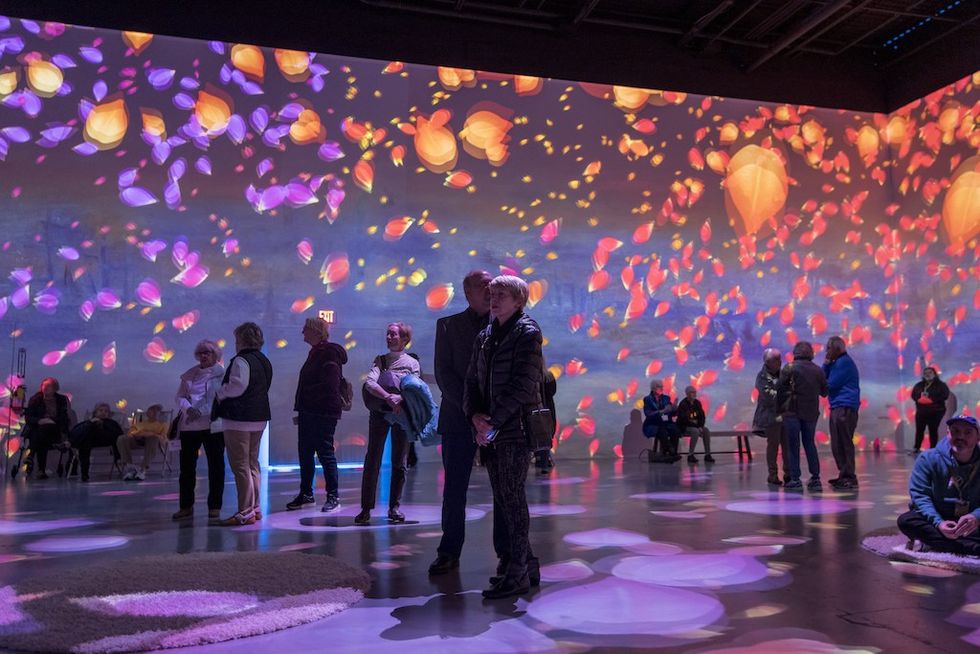 People in immersive art space with vibrant floral projections on walls.